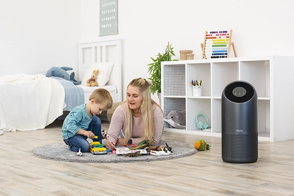 A young woman is lying on the floor in the children's room with a child and playing. Next to it is the Hama "Smart" air purifier