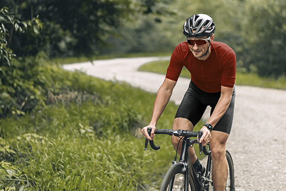 Ein Mann sitzt in kurzer Radlerkleidung auf einem Rennrad und radelt einen geschotterten Waldweg entlang. Am Handgelenk trägt er eine Hama Smartwatch.