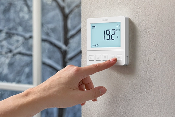 Das smarte Hama Fußbodenheizungs-Thermostat ist an einer Wand neben einem Fenster befestigigt und wird von einer Hand bedient.