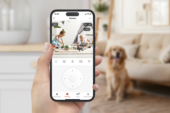 Im Vordergrund sieht man eine Hand, die ein Smartphone mit geöffneter Smart Home-App hält. Auf der Videoübertragung der App sind zwei Kleinkinder beim Spielen zu sehen. Im Hintergrund des Bildes ist unschaft ein Wohnzimmer mit Hund zu sehen.