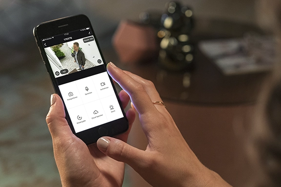 You can see a woman holding a smartphone with the Smart Home app open. The display shows a woman in front of an entrance door.