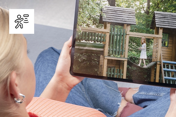 A woman sits on the sofa and looks at a tablet in her hands. It shows a young girl playing on a wooden climbing frame.