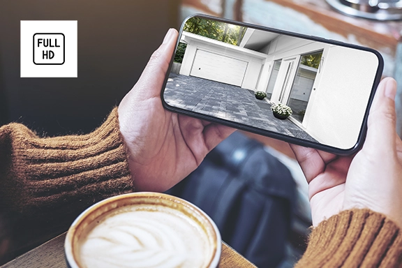 Someone sits in a café with a cup of cappuccino in front of them and looks into their smartphone, where they can see a driveway, front door and garage. The image is transmitted by the Hama smart surveillance camera.