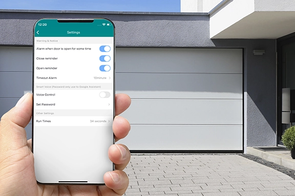 Im Vordergrund hält eine Hand ein Smartphone mit geöffneter Smart Home App. Im Hintergrund sieht man eine Hofeinfahrt mit geschlossenem Garagentor.