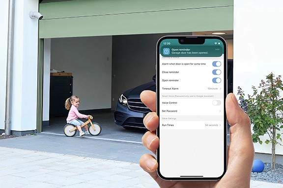 Im Vordergrund hält eine Hand ein Smartphone mit geöffneter Smart Home App. Im Hintergrund sieht man eine Hofeinfahrt mit geöffnetem Garagentor.